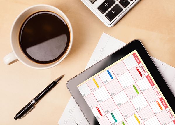 A scheduling tablet with a calendar displayed sits next to a cup of coffee.